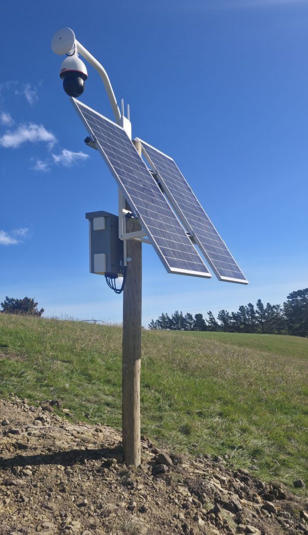 Solar-powered security camera with panels on pole in rural field under blue sky